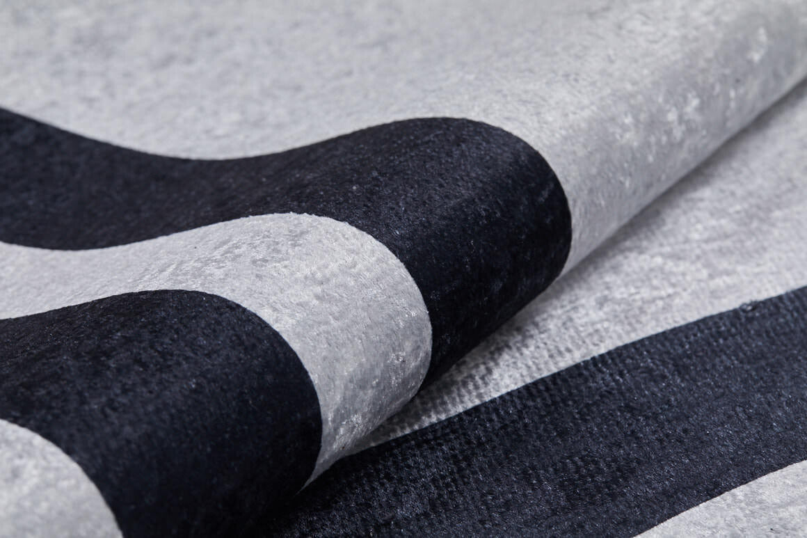 Close-up of the Timeless Border - Washable Rug - JR572 (Custom Size) in gray-anthracite with bold, curved black stripes. Its soft, subtly shiny material is partially folded—durable and washable for high-traffic areas.