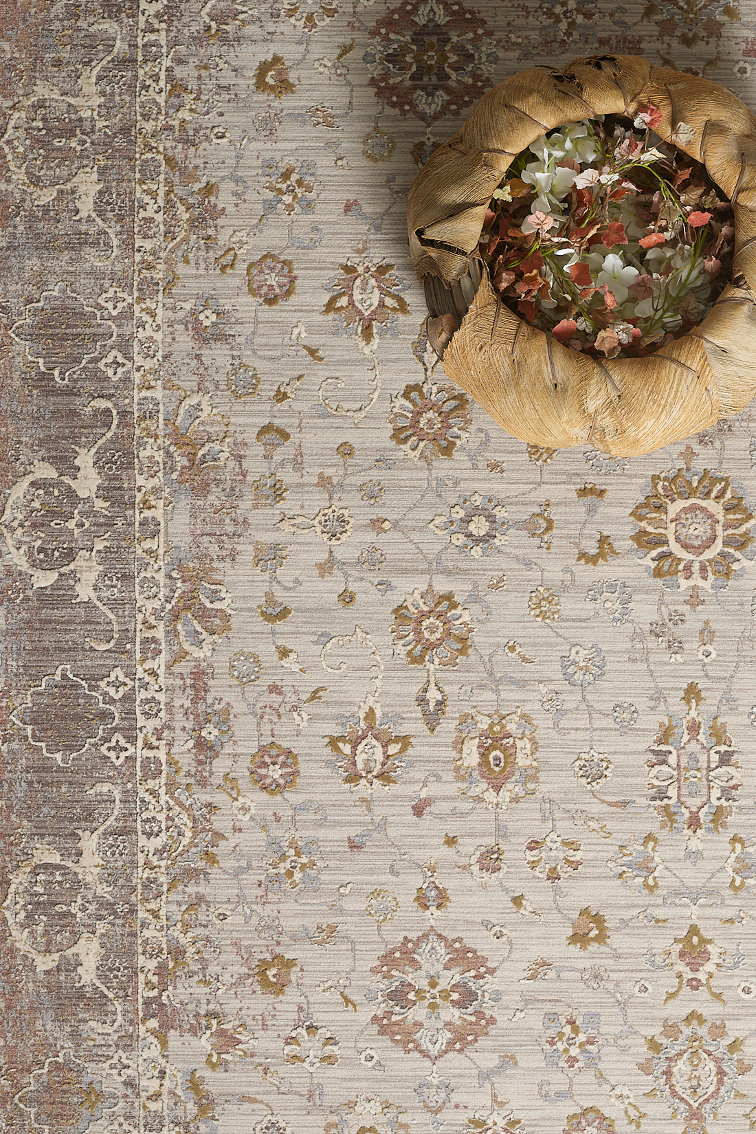 A decorative woven basket with pink and white flowers sits on the Timeless Petals Oushak Rug - Sand - M621N, a beige and taupe patterned, machine-made area rug with intricate floral and geometric designs.