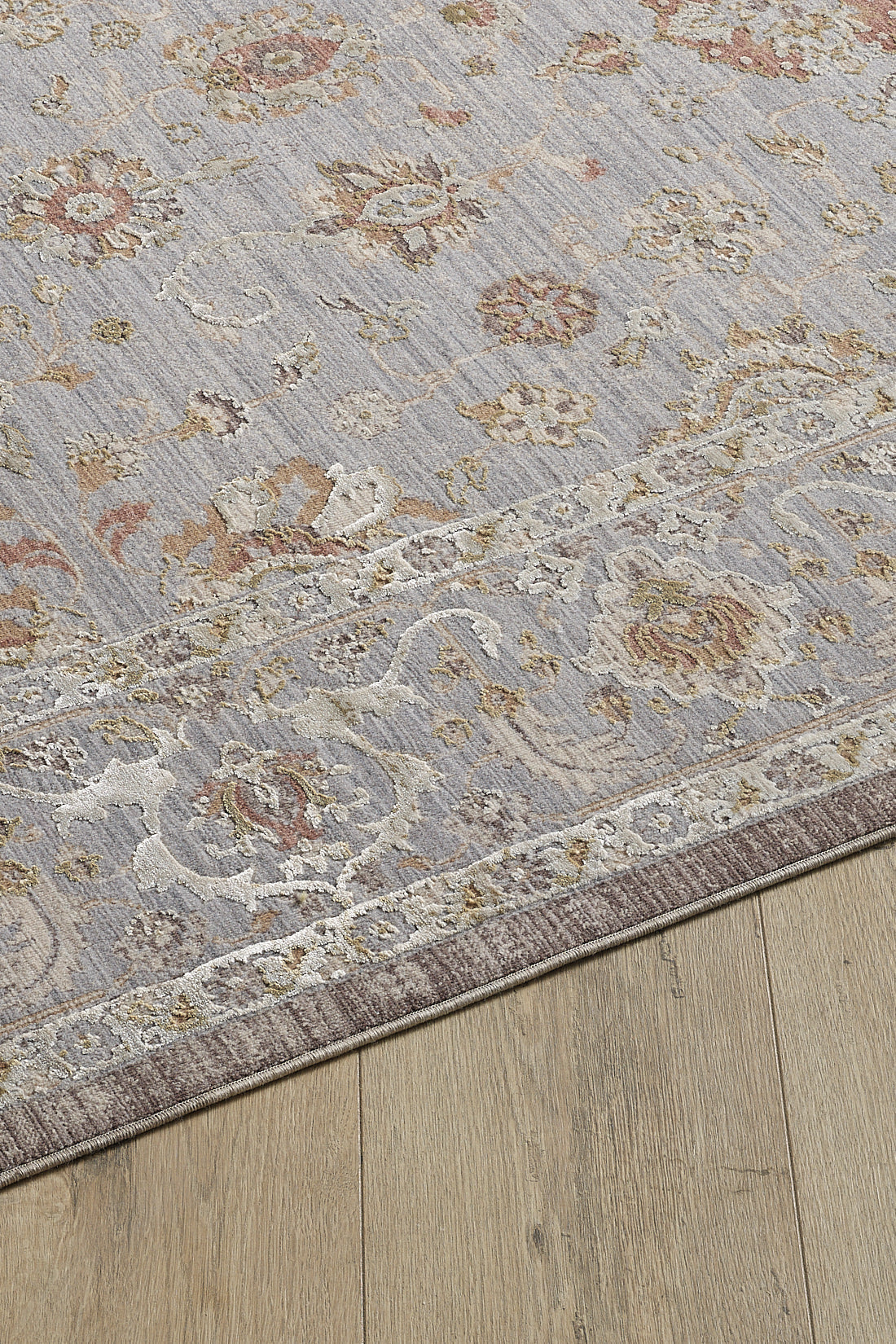 A close-up of the Timeless Petals Oushak Rug - Silver - M621O, featuring a floral and vine pattern in muted pastels with a beige border, placed on a light wood floor.