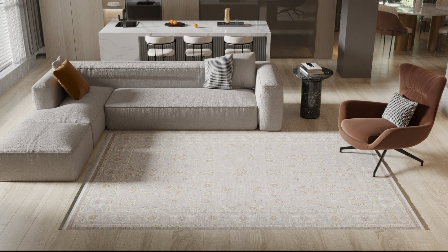 A modern living room features a light gray sectional, brown lounge chair, marble side table, kitchen island with barstools in the background, and is anchored by the Timeless Petals Oushak Rug - Silver - M621O. Natural light fills the space.