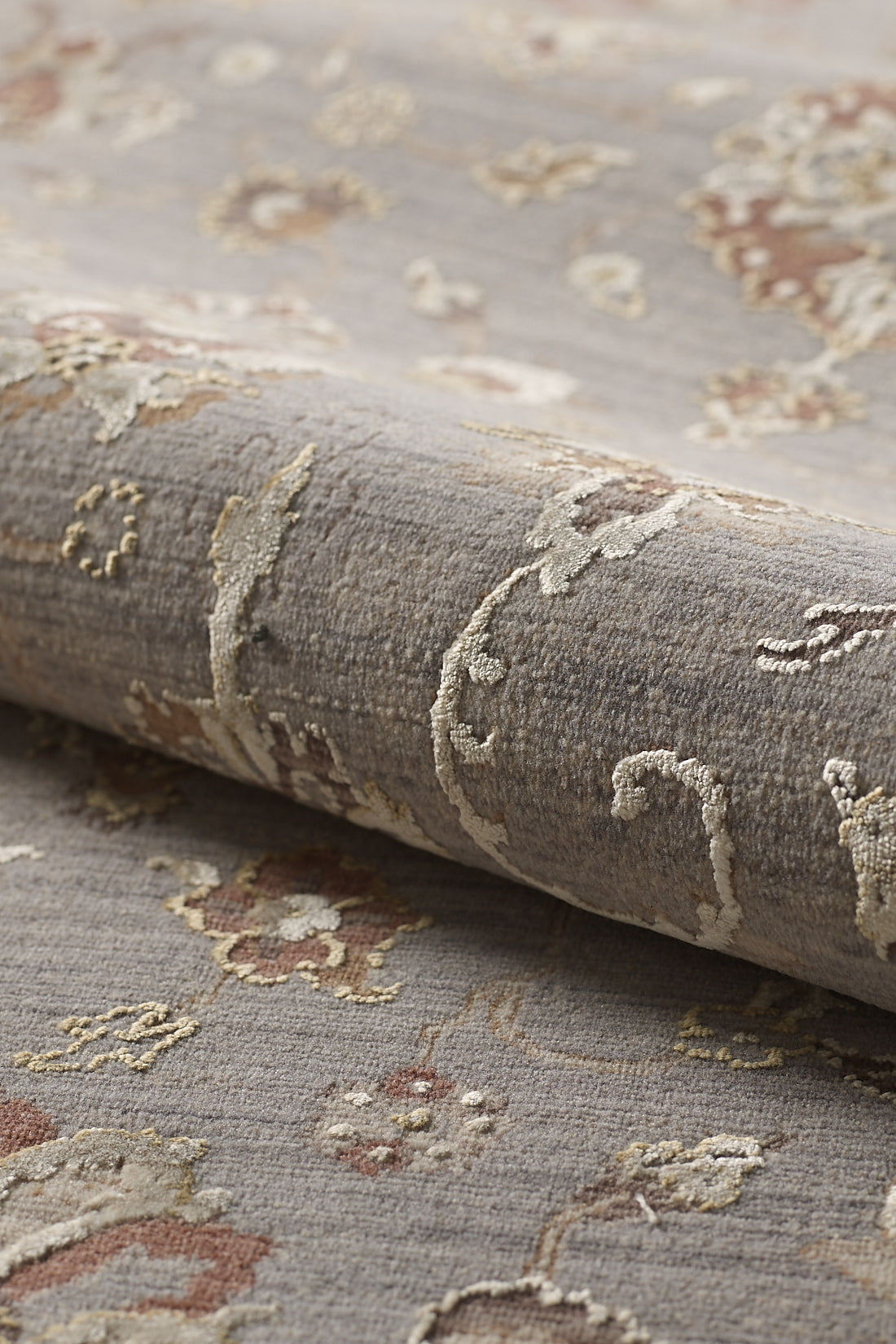 A close-up of the Timeless Petals Oushak Rug - Silver (M621O) shows its textured beige and light brown pattern on a muted gray background, with a soft surface and slightly raised areas for added depth.