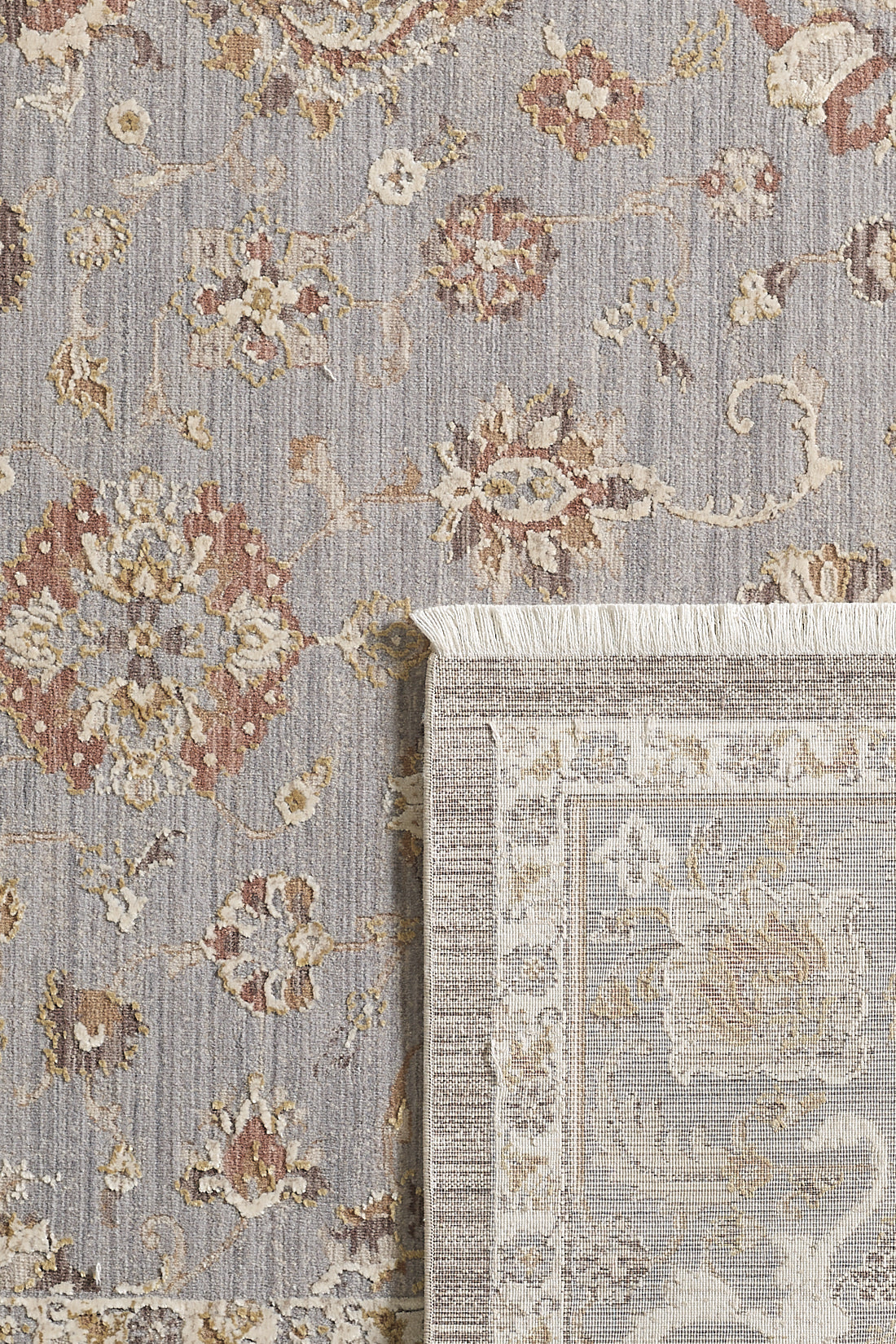 A close-up of the Timeless Petals Oushak Rug - Silver - M621O shows muted beige, gray, and brown floral and vine patterns. A flipped corner reveals the backside and fringe edge, highlighting its quality craftsmanship.