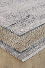 A close-up of the Timeless Remix Bordered Rug - Beige - MT187, a machine-made rug with a distressed gray pattern, gold and gray Greek key border, and short white fringe, seen on a light wooden floor.
