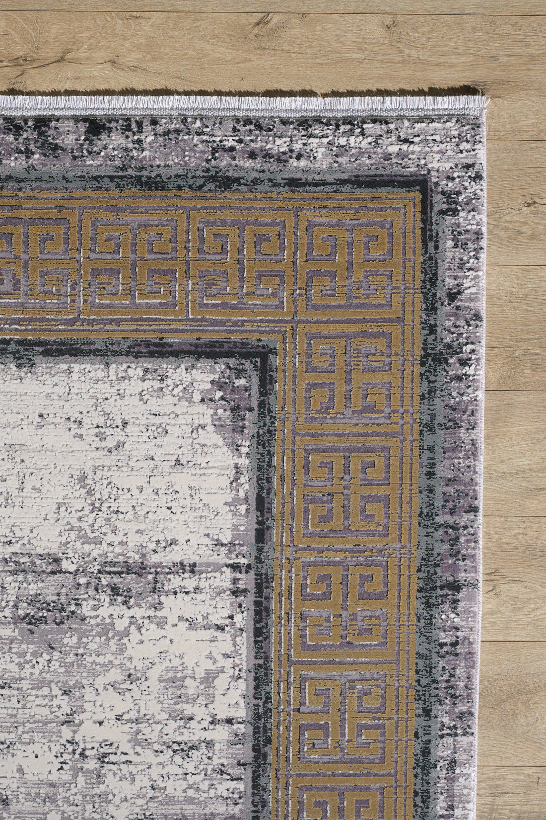 A close-up of the Timeless Remix Bordered Rug - Beige - MT187 on a wooden floor, featuring a gold and purple Greek key border and a distressed abstract gray and white main area.