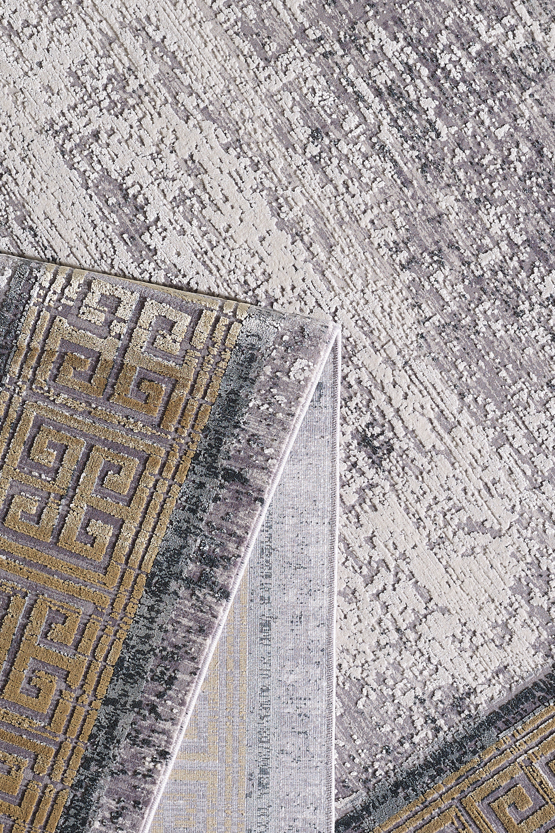 A close-up of the Timeless Remix Bordered Rug - Beige (MT187) shows a folded corner revealing its geometric Greek key pattern in gold and gray on a mottled light gray and cream background.
