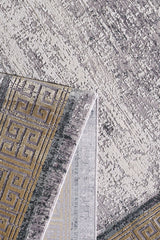 A close-up of the Timeless Remix Bordered Rug - Beige (MT187) shows a folded corner revealing its geometric Greek key pattern in gold and gray on a mottled light gray and cream background.