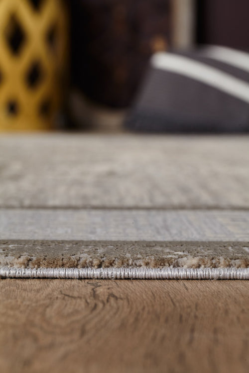 Load image 1 in gallery view. Close-up side view of the edge of the Timeless Remix Bordered Rug - Cream - MT189 on a wooden floor, with blurred yellow patterned decor and a black-and-white striped pillow in the background.
