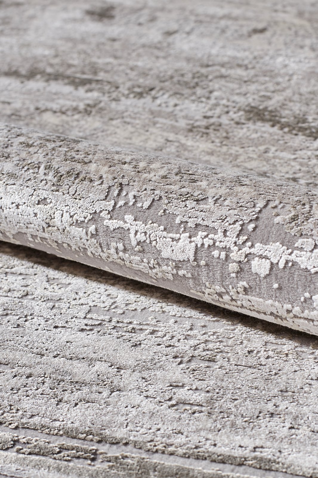A close-up of the Timeless Remix Bordered Rug - Cream - MT189, rolled up to show its textured grey and beige acrylic-viscose surface with an abstract, weathered pattern and a soft, plush feel.