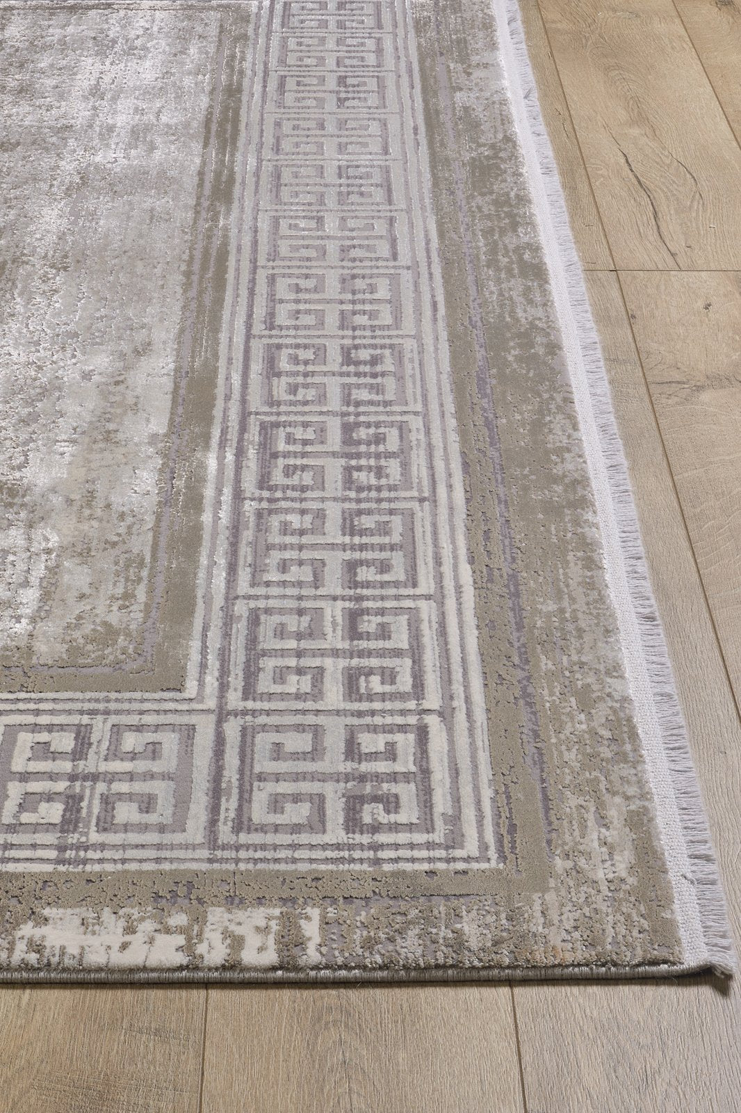 A close-up of the Timeless Remix Bordered Rug - Cream - MT189, a rectangular machine-made acrylic-viscose rug with a light gray and beige Greek key border and a slightly distressed, textured look, shown on a wooden floor.