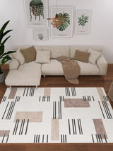 A modern living room featuring the Timeless Scroll - Washable Rug - JRN45, a beige sectional, brown and beige cushions, a tan throw, green plants, wall art—ideal for high-traffic contemporary spaces with its black and brown geometric pattern.