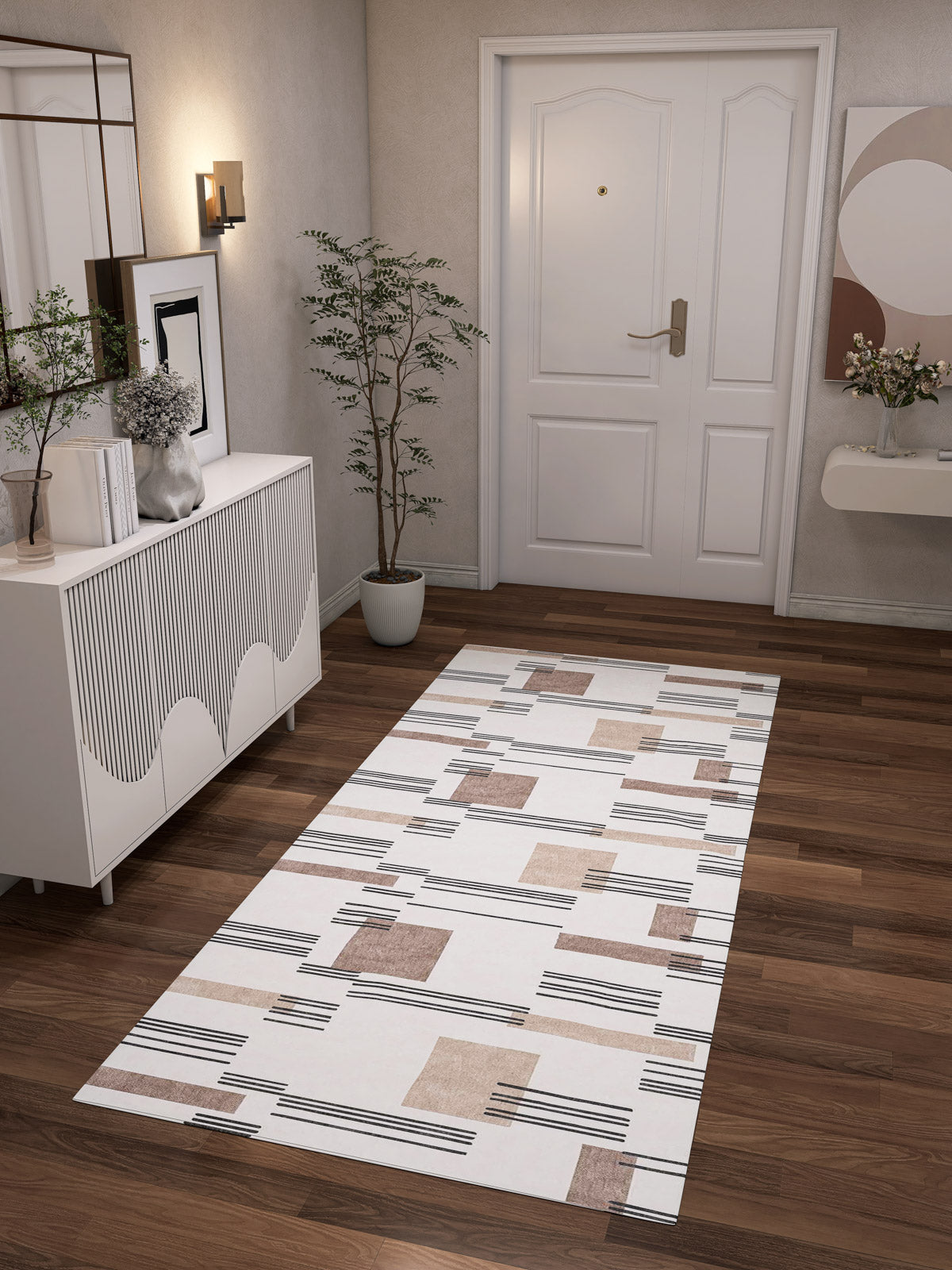 A modern hallway with a dark wooden floor, white door, and white furniture features the Timeless Scroll - Washable Rug - JRN45. Decorative plants, wall art, a mirror, and soft lighting complete this cozy space—ideal for high-traffic areas.