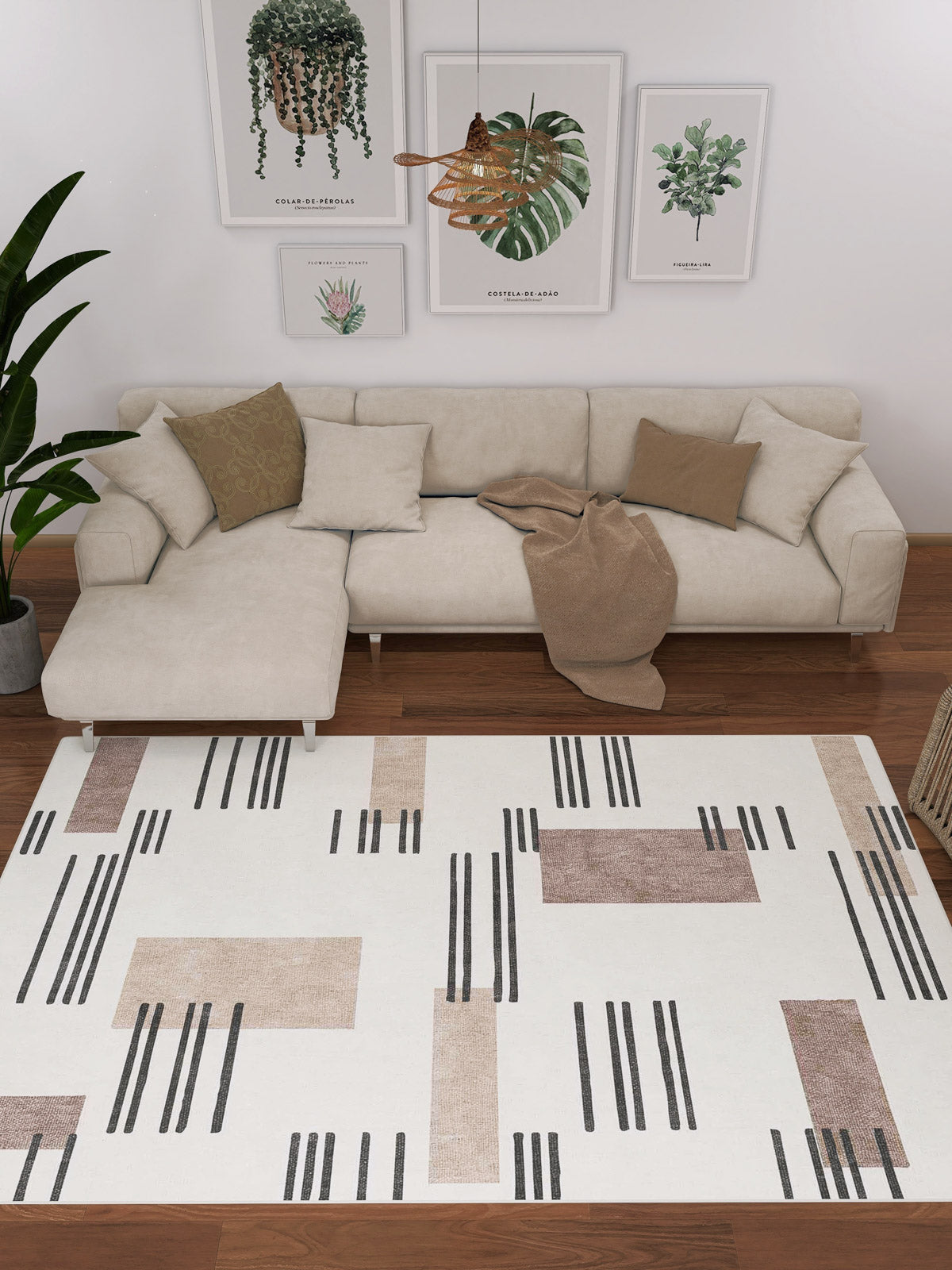 A contemporary living room features a beige sectional, earth-toned cushions, and a tan throw. The custom-size Timeless Scroll - Washable Rug - JRN45 with geometric patterns covers the floor, while framed botanicals and a hanging plant adorn the wall.