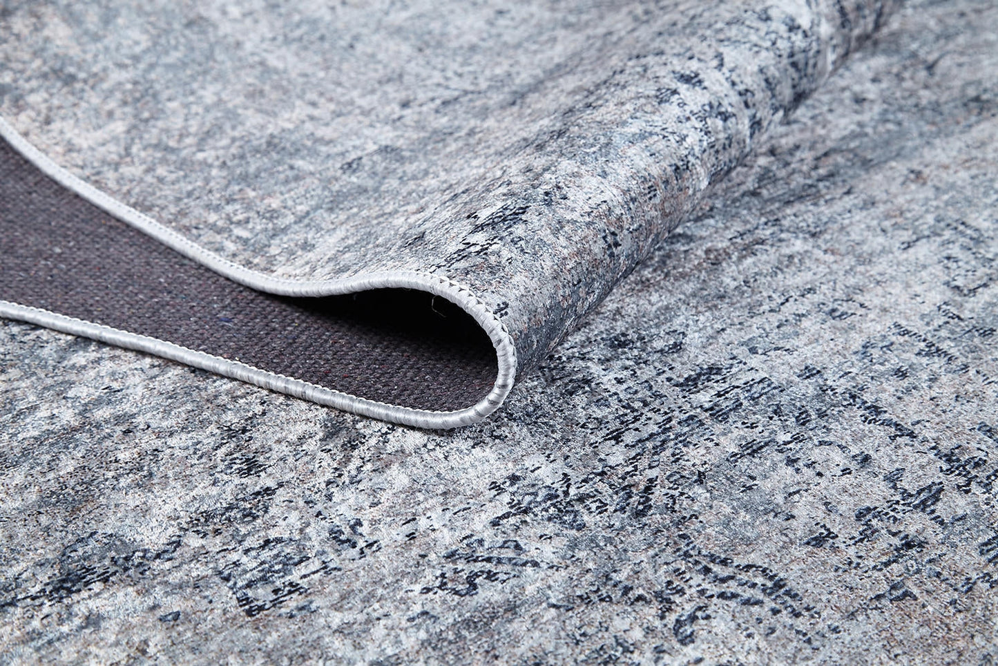 A close-up of the Timeless Scrollwork - Washable Rug - JR1774 (Custom Size) shows its textured gray and blue abstract pattern with a corner folded to display the dark, non-slip backing—perfect for high-traffic areas.