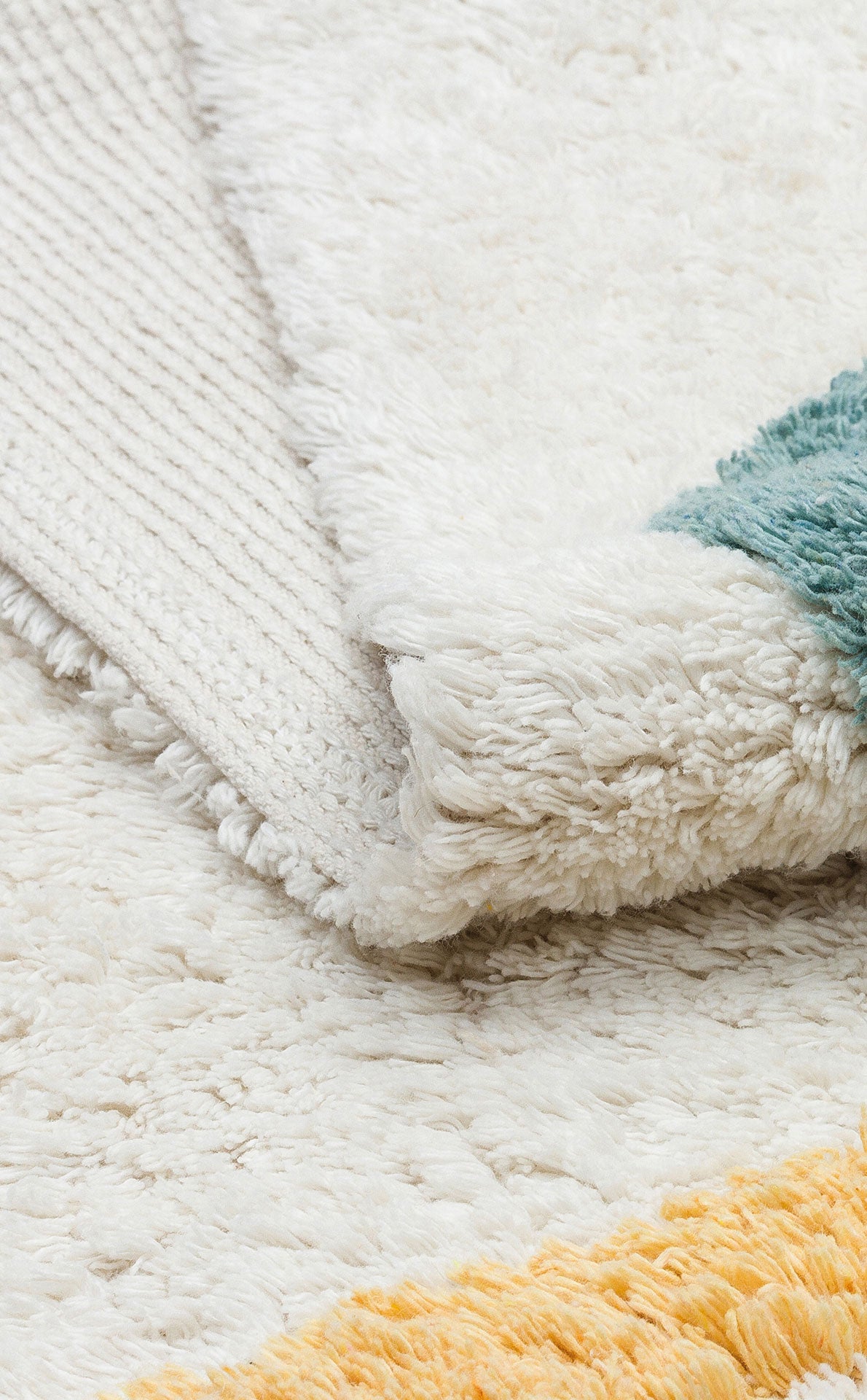 Close-up of the Tiny Joy - Washable Shaggy - CBN02 kids room rug, featuring a soft white pile with teal and golden yellow tufted edges. A folded corner reveals its textured underside.