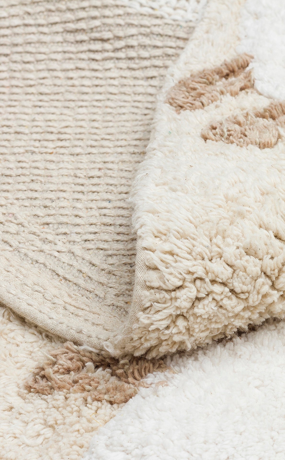 A close-up of the Tiny Motion - Washable Shaggy - CBN11 rug, folded to reveal its cream-colored, cotton texture with brown spots, highlighting both the soft shaggy surface and the woven underside.