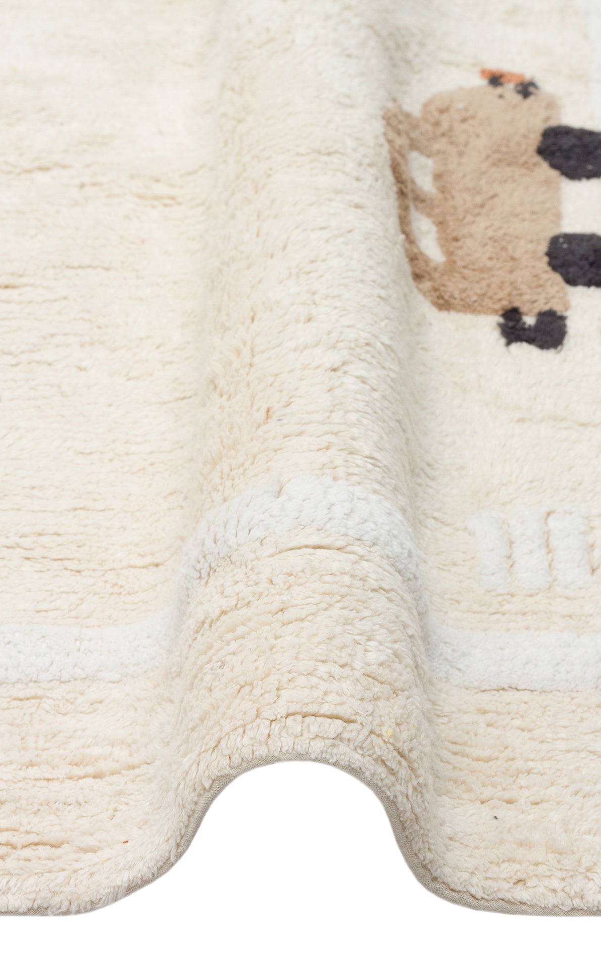 Close-up of the Tiny Voyage - Washable Shaggy - CBN09 cream cotton playroom rug, showcasing its textured surface and small brown and black animal design with a wavy, folded edge against a white background.