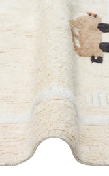 Close-up of the Tiny Voyage - Washable Shaggy - CBN09 cream cotton playroom rug, showcasing its textured surface and small brown and black animal design with a wavy, folded edge against a white background.