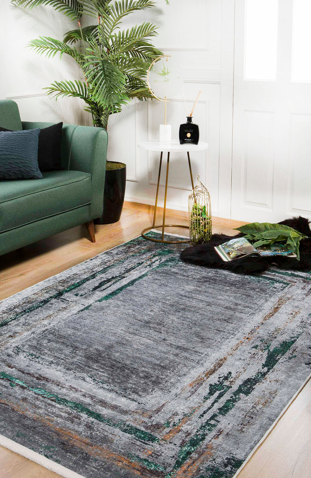 A modern living room with a green sofa, the JR1543 Traditional and Classic Washable Rug in gray, a round white side table with gold legs, potted plant, and decor. Light wood floors and white walls provide a bright, airy backdrop.
