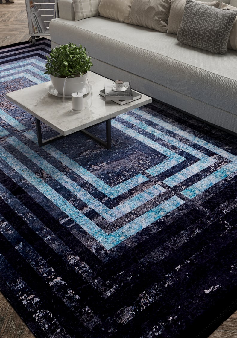A modern living room with a white sofa, gray and white cushions, a marble coffee table, and the Traditional and Timeless - Washable Rug - JR1408—a blue geometric rug perfect for high-traffic areas. A potted plant and tray with cups rest on the table.
