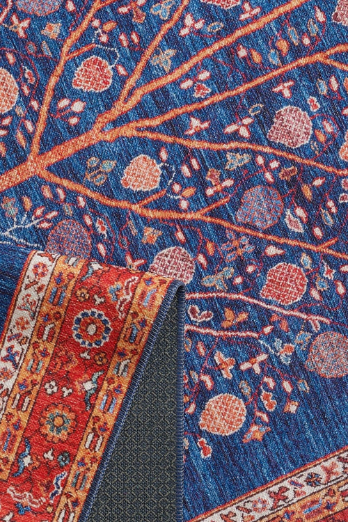Load image 1 in gallery view. The Tree of Plenty Red Blue Washable Rug (LCC3031) features a blue background with tree branches and pomegranates, plus a red patterned border. A folded corner reveals the underside—perfect for high-traffic use.