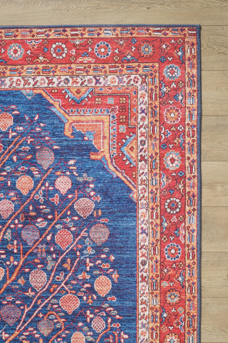 The Tree of Plenty Red Blue Washable Rug - LCC3031 features a vibrant red and orange geometric border, blue background with floral and tree motifs, ideal for allergy-sensitive homes and high-traffic areas, shown on a light wood floor.