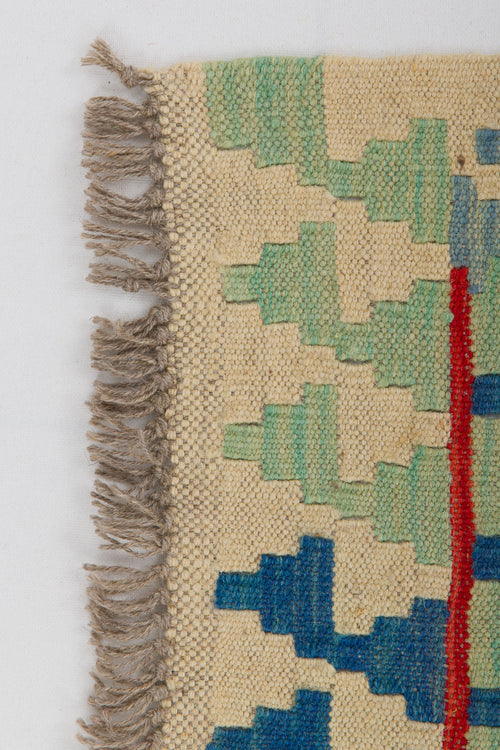 Load image 1 in gallery view. A close-up of the Tribal Essence - Handwoven Kilim shows beige, green, and blue geometric patterns, a red vertical stripe, and brown fringes on the corner, all set against a white background.