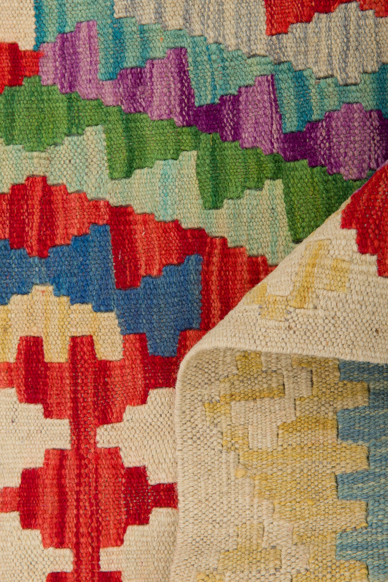 The Tribal Essence - Handwoven Kilim features colorful geometric patterns in red, blue, green, and purple with a folded corner revealing its lighter underside—an ideal choice for kilim enthusiasts seeking vibrant flatweave décor.