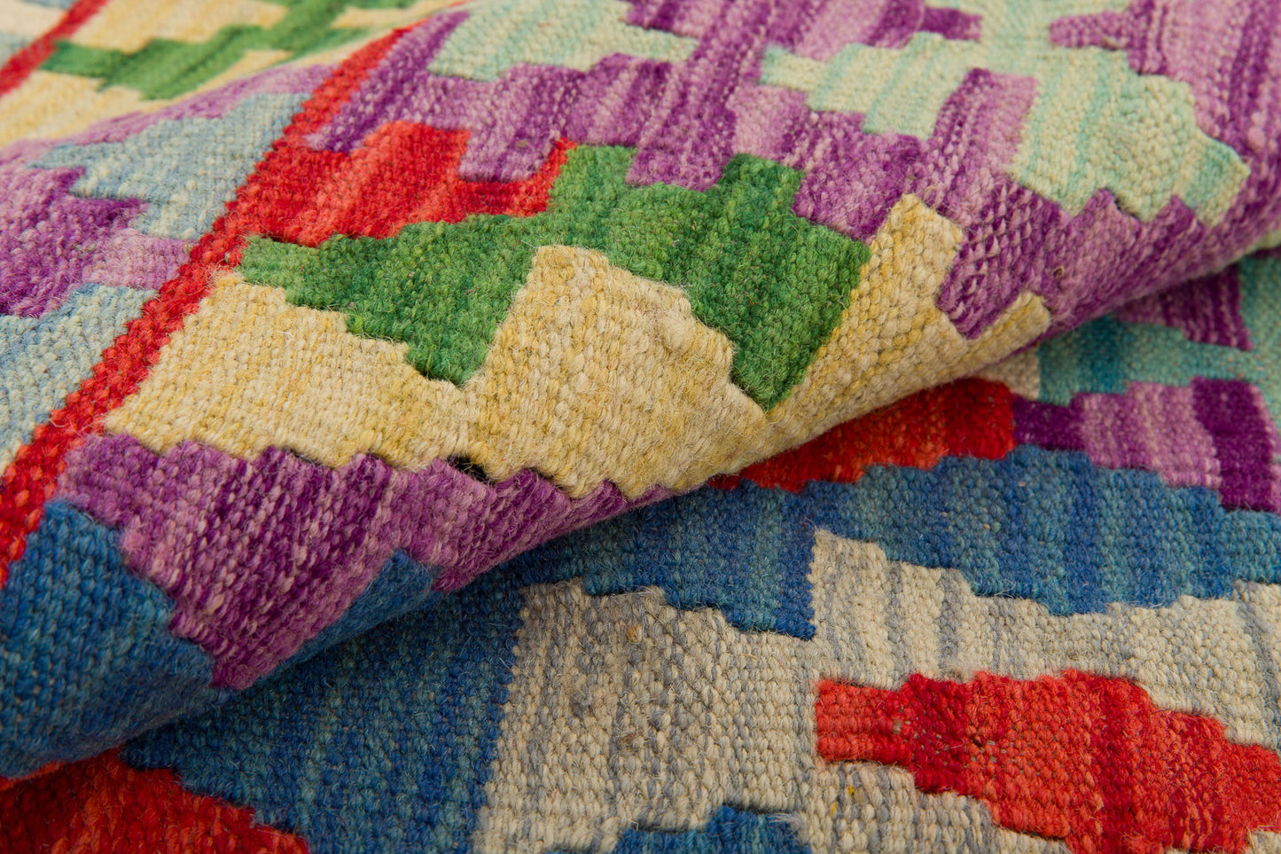 A close-up of Tribal Essence - Handwoven Kilim showcases vivid geometric patterns in purple, green, red, yellow, and blue. The folded wool fabric reveals sharp zigzag shapes and intricate designs typical of a traditional kilim rug.