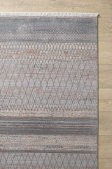 The Tribal Essence Trellis Rug - HRD005 features a modern geometric diamond pattern in muted gray and blue with touches of rust and beige. Machine-made, it has white fringes on one edge and is shown on a light wood floor.