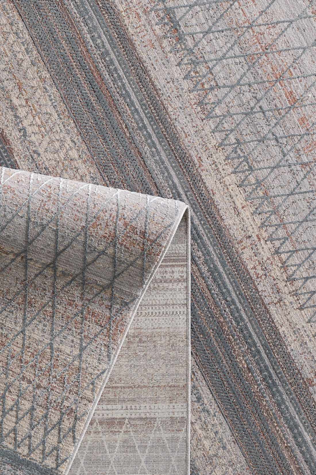 A close-up of the Tribal Essence Trellis Rug - HRD005, showcasing its folded corner and geometric patterns in muted gray, beige, and rusty brown. This machine-made rug combines durability with contemporary style.