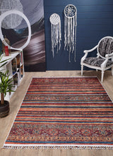 The Tropic Thunder - Washable Rug - JR5077 (Custom Size) adds color to a modern room with a wooden floor, white chair, small bookshelf, potted plant, and two white dreamcatchers on a dark blue wall.