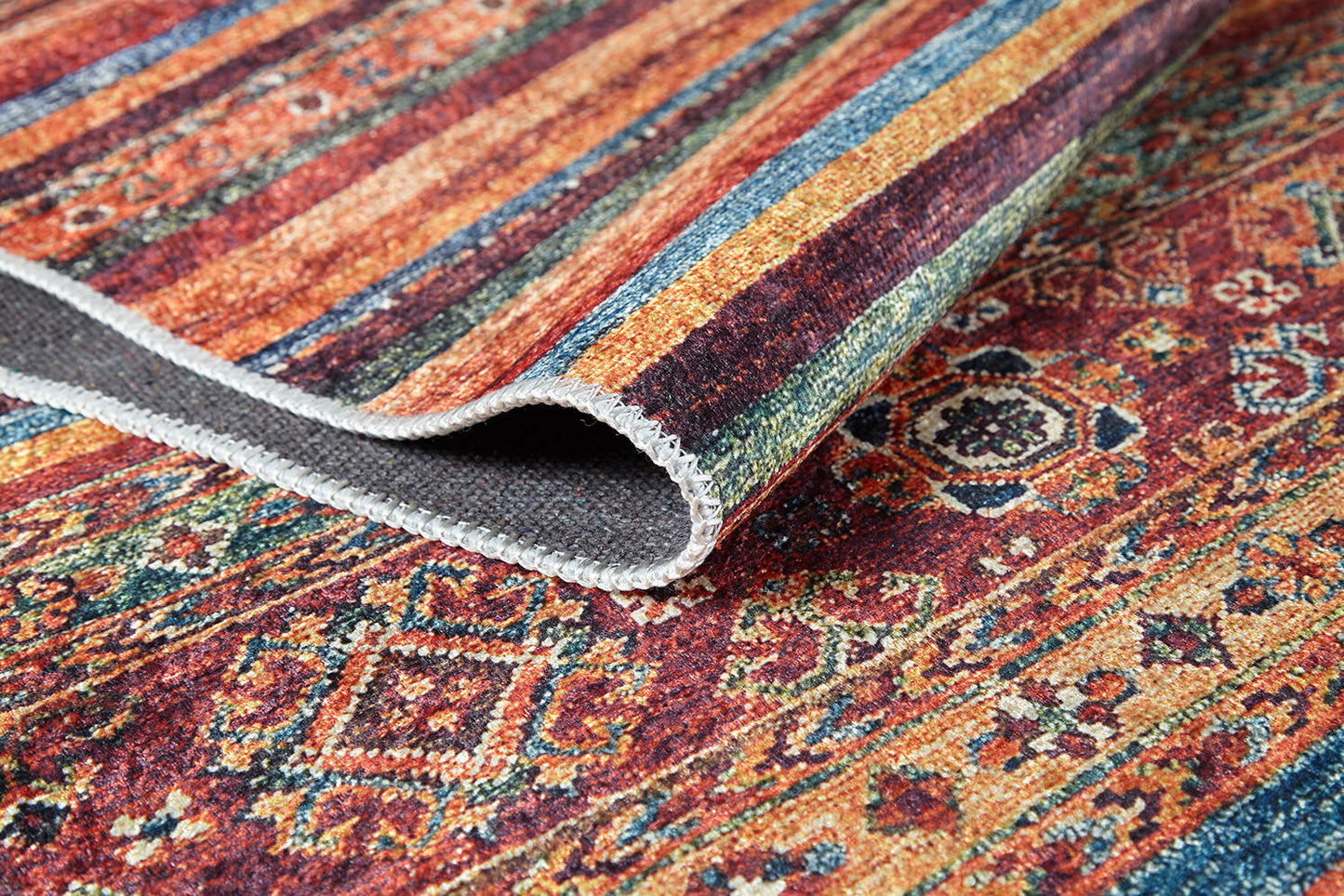 A close-up of the Tropic Thunder - Washable Rug - JR5077 (Custom Size), featuring vibrant geometric and floral patterns in red, orange, blue, and green. One corner is folded to reveal its textured, allergy-friendly underside.