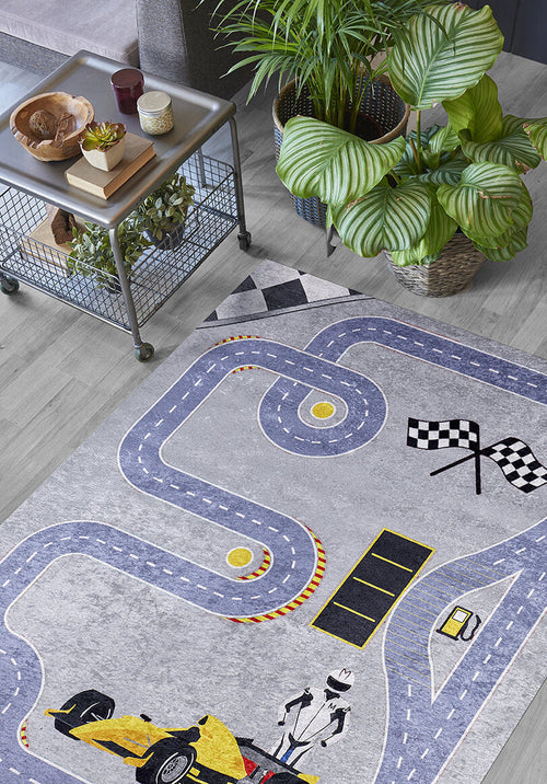 Load image 1 in gallery view. The Turbo Tracks - Machine-Washable Rug JRC089 (Custom Size) with a racetrack, finish line, and race car design is displayed on a light wood floor beside a metal cart with wooden bowls and potted plants, plus large leafy plants in the background.