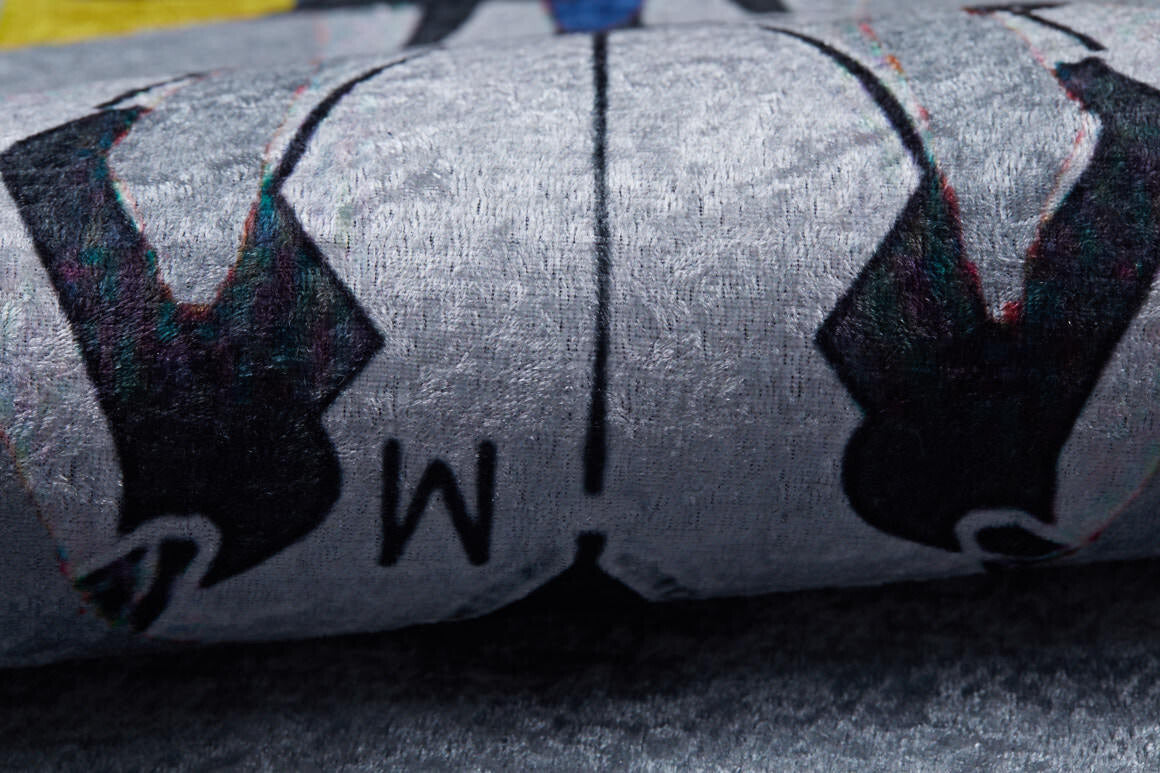 Close-up of the Turbo Tracks - Machine-Washable Rug JRC089 (Custom Size) in gray-anthracite, showing its flat weave, black shapes, blue accent, and central "M" design. The washable rug appears slightly rolled or folded.