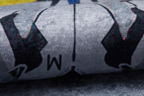Load image 1 in gallery view. Close-up of the Turbo Tracks - Machine-Washable Rug JRC089 (Custom Size) in gray-anthracite, showing its flat weave, black shapes, blue accent, and central "M" design. The washable rug appears slightly rolled or folded.