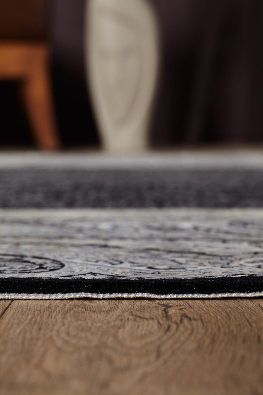 Close-up view of the Turkish Mirage Silk Medallion Rug - Obisidan - 1168D with a paisley border on a wooden floor; in the blurred background are a brown chair and an abstract, neutral-toned décor piece.