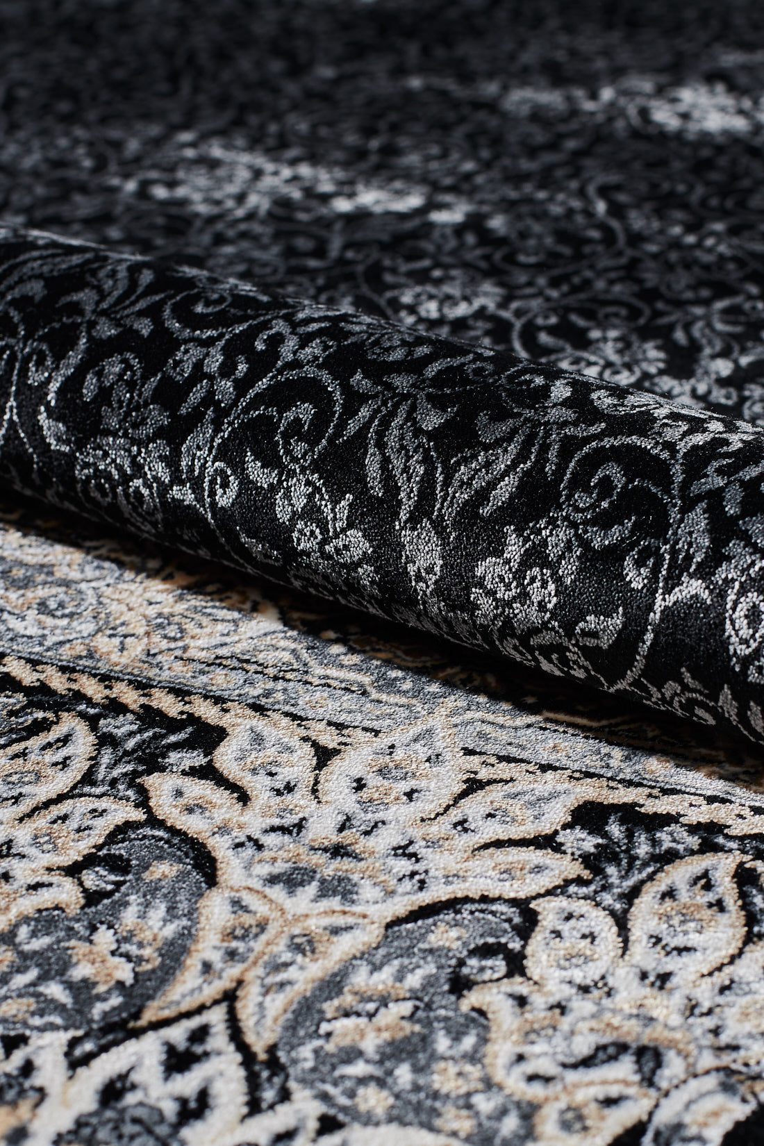 A close-up of the Turkish Mirage Silk Medallion Rug in Obsidian (1168D) being partially rolled up, highlighting its ornate black, gray, and cream swirling designs with a subtle paisley border.