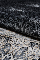 A close-up of the Turkish Mirage Silk Medallion Rug in Obsidian (1168D) being partially rolled up, highlighting its ornate black, gray, and cream swirling designs with a subtle paisley border.
