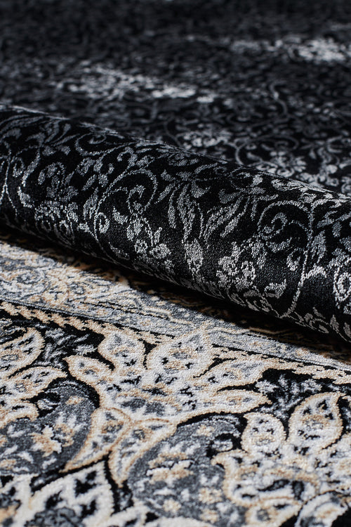 Load image 1 in gallery view. A close-up of the Turkish Mirage Silk Medallion Rug in Obsidian (1168D) being partially rolled up, highlighting its ornate black, gray, and cream swirling designs with a subtle paisley border.