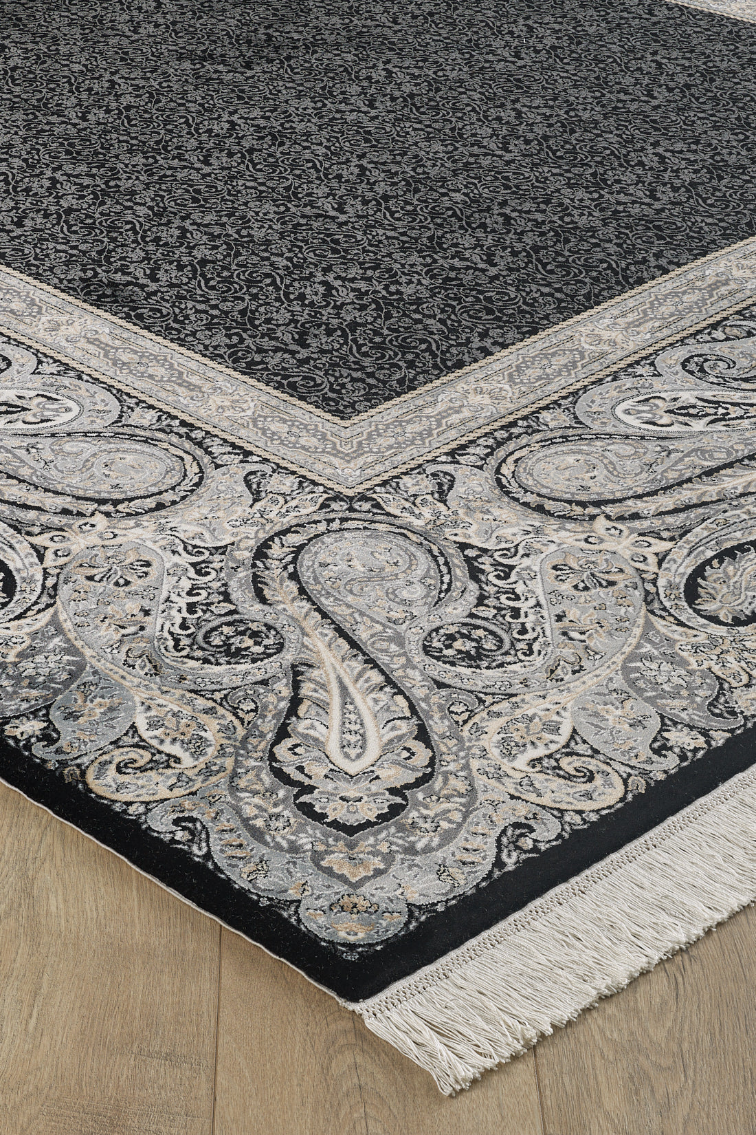 A close-up of the Turkish Mirage Silk Medallion Rug - Obisidan - 1168D, a rectangular modal silk rug with black, gray, and cream paisley patterns, ornate borders, and white fringe on one edge, shown on a light wood floor.