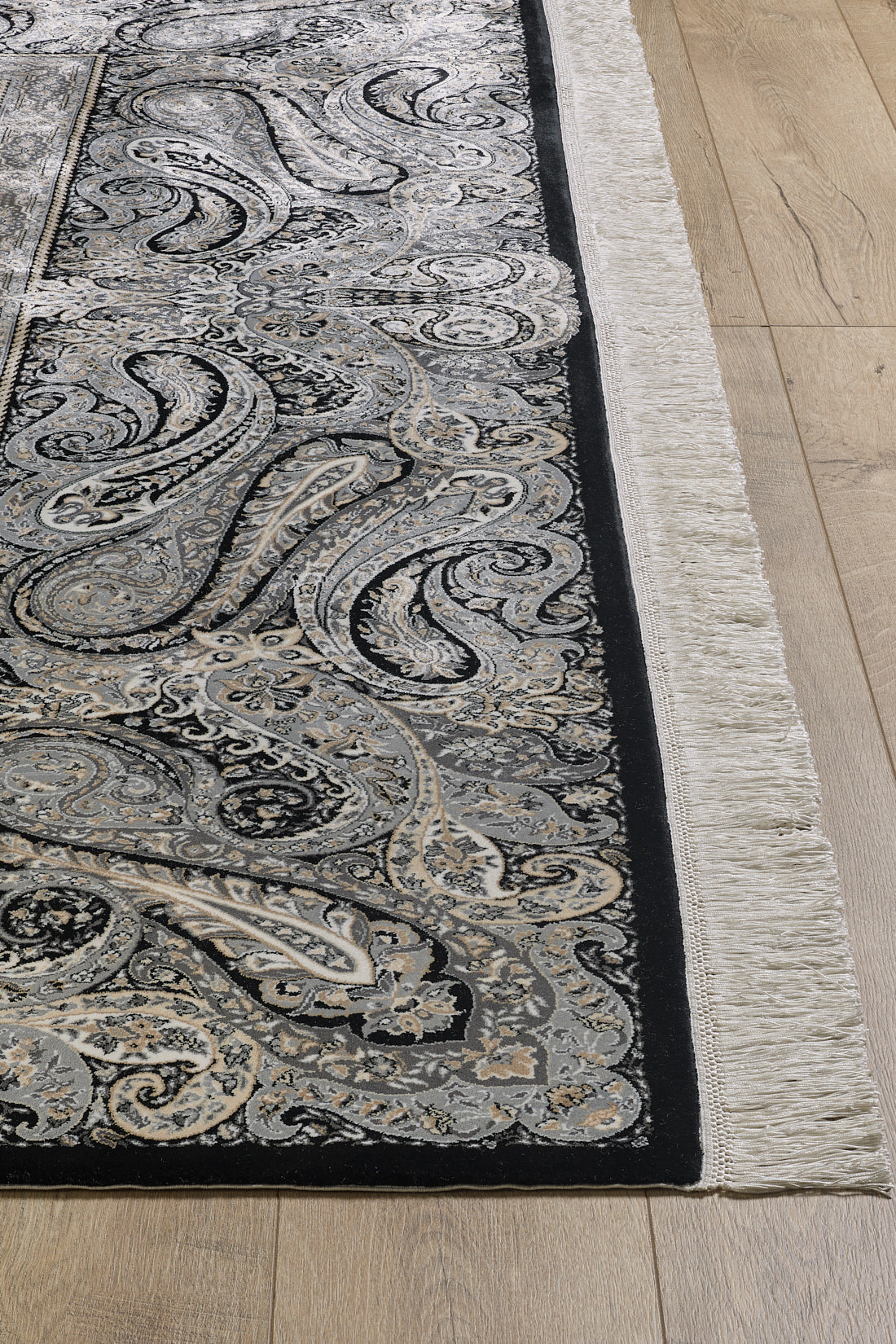 A close-up of the Turkish Mirage Silk Medallion Rug - Obisidan - 1168D showcasing its intricate paisley and floral patterns in black, gray, and cream with white fringed edges on a light wood floor.