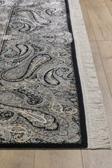 A close-up of the Turkish Mirage Silk Medallion Rug - Obisidan - 1168D showcasing its intricate paisley and floral patterns in black, gray, and cream with white fringed edges on a light wood floor.