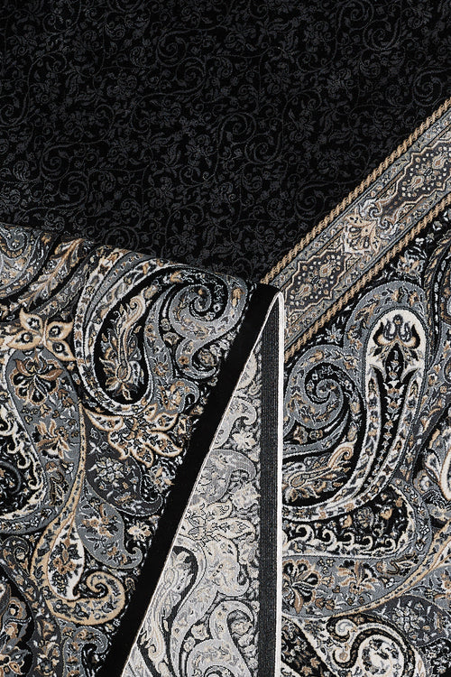 Load image 1 in gallery view. A close-up of the Turkish Mirage Silk Medallion Rug - Obsidian - 1168D showcases intricate paisley and floral patterns in black, gray, and beige, with a section folded to reveal its contrasting underside design.