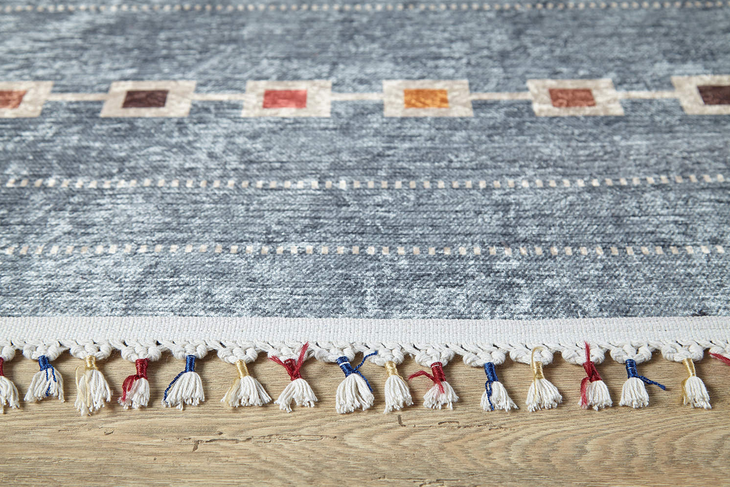 A close-up of the Tuscan Villa - Washable Rug - JR5058 (Custom Size) in blue, featuring textured fabric, a row of colored edge squares, and white tassels with red and blue accents—perfect for high-traffic spaces on a wooden floor.