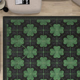 A top-down view of the Clover Noir - Washable Rug - JRS012, featuring a repeating green four-leaf clover pattern on a dark background, perfect for high-traffic areas, placed on light wood flooring near light chairs and an open magazine.