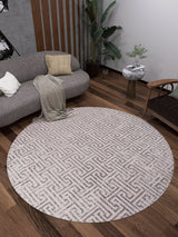 A modern living room with wooden floors showcases the Gatsby Glam - Washable Rug - JR1223 (Outlet) US in round cream and beige, paired with a gray curved sofa, pillows, throw, wooden armchair, side table with plant, and a tall green plant in the corner.