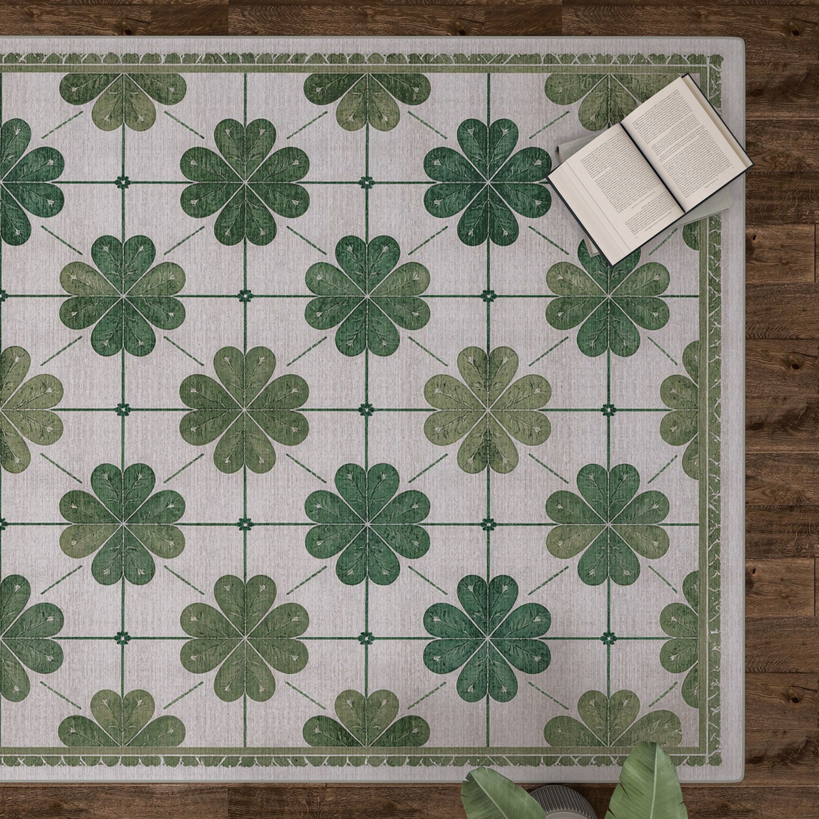 The Emerald Charm - Washable Rug - JRS013, featuring green four-leaf clovers and a subtle floral pattern, lies on a wooden floor with books and plant leaves nearby—ideal for high-traffic areas.