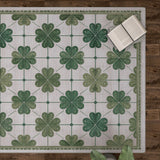 The Emerald Charm - Washable Rug - JRS013, featuring green four-leaf clovers and a subtle floral pattern, lies on a wooden floor with books and plant leaves nearby—ideal for high-traffic areas.
