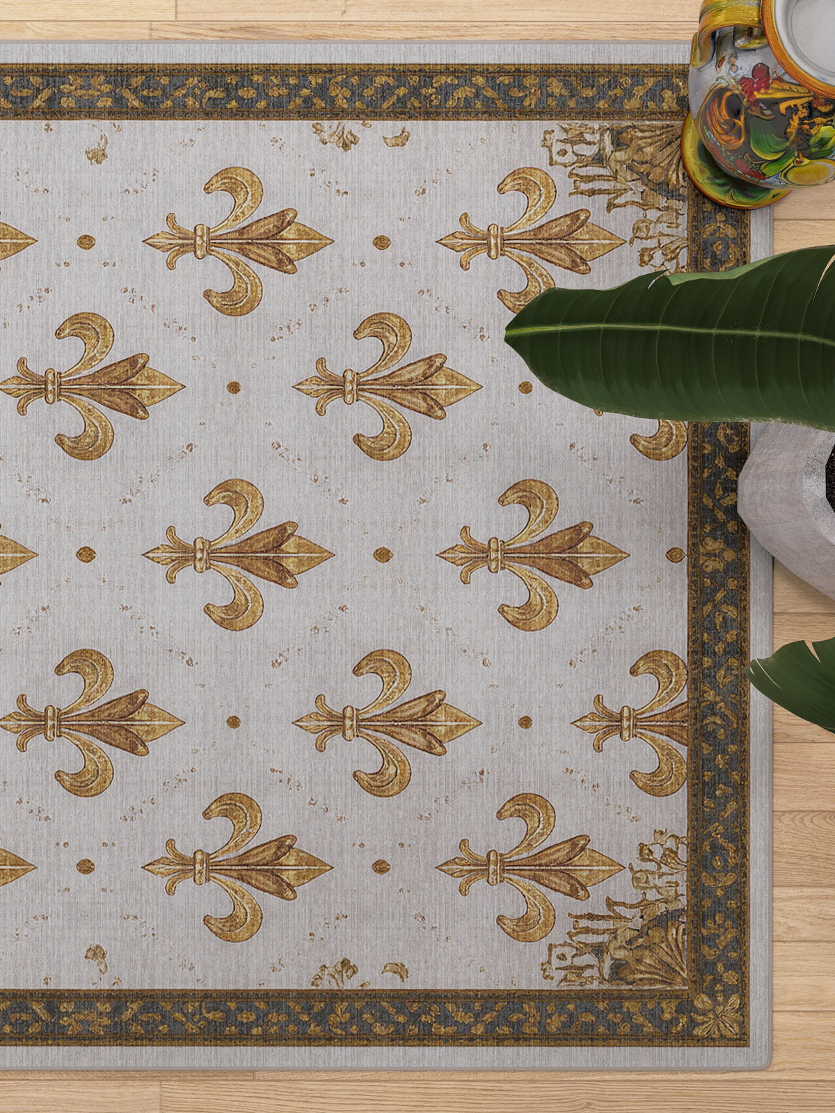The Golden Crest - Washable Rug (JRS015) features gold fleur-de-lis on a light background with a dark and gold ornate border—ideal for allergy-sensitive or high-traffic spaces. A leafy green plant in a colorful pot appears at the top right corner.