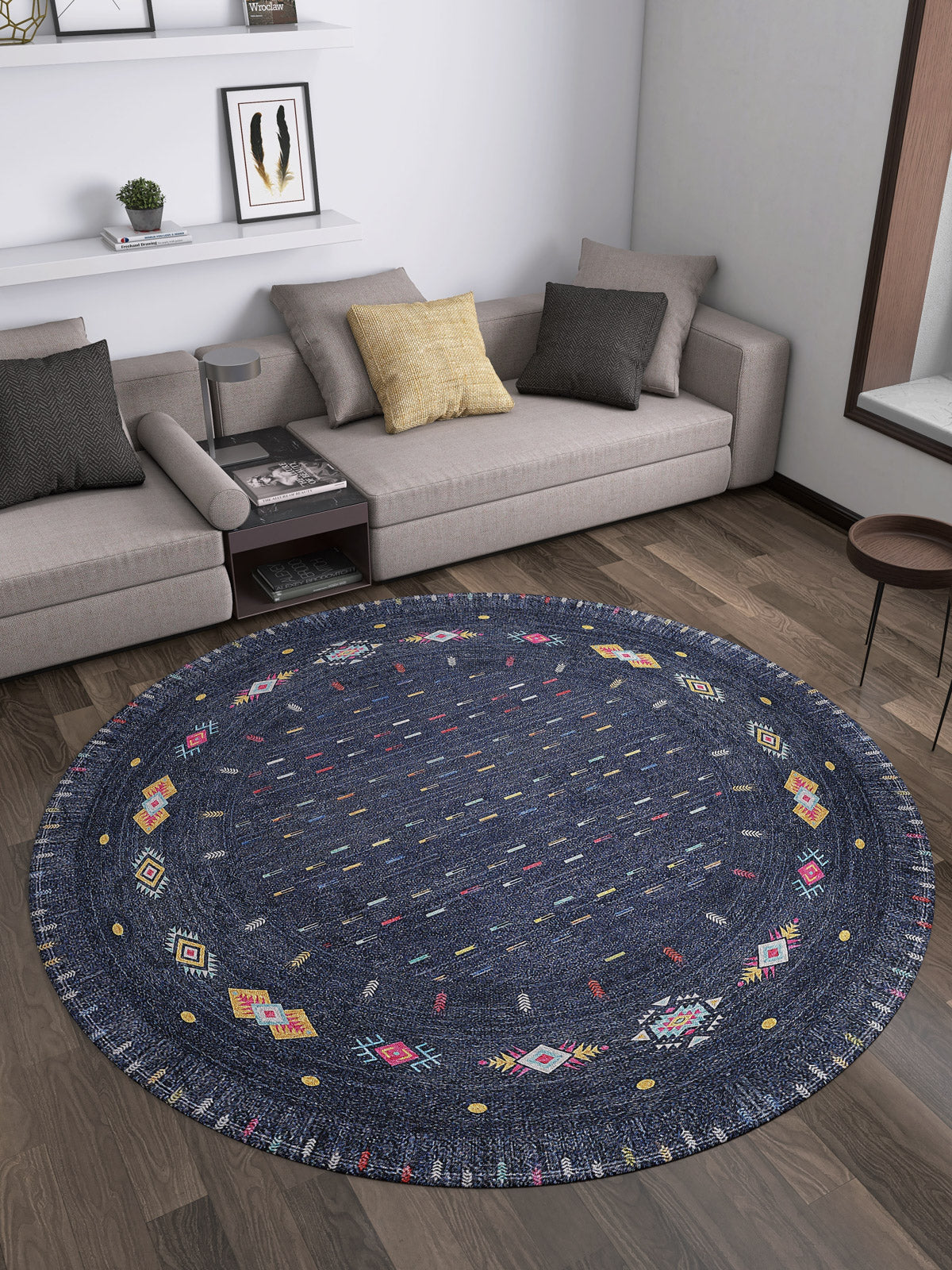 A modern living room with the Wanderer's Way - Washable Rug - JR5101 in blue, featuring colorful geometric patterns, a gray sectional sofa, assorted cushions, a round table, and wall shelves—ideal for high-traffic spaces.
